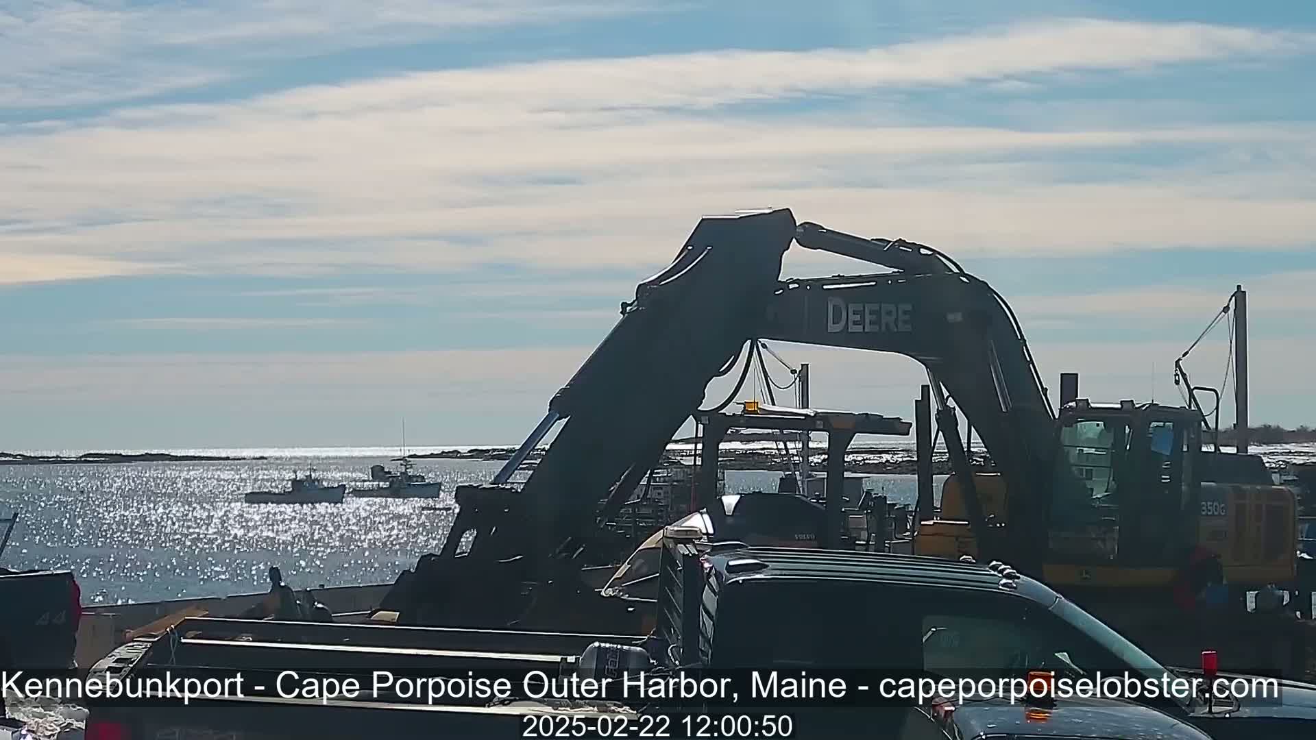 A large excavator sits near several fishing boats in a harbor on a sunny day with a mostly clear sky.