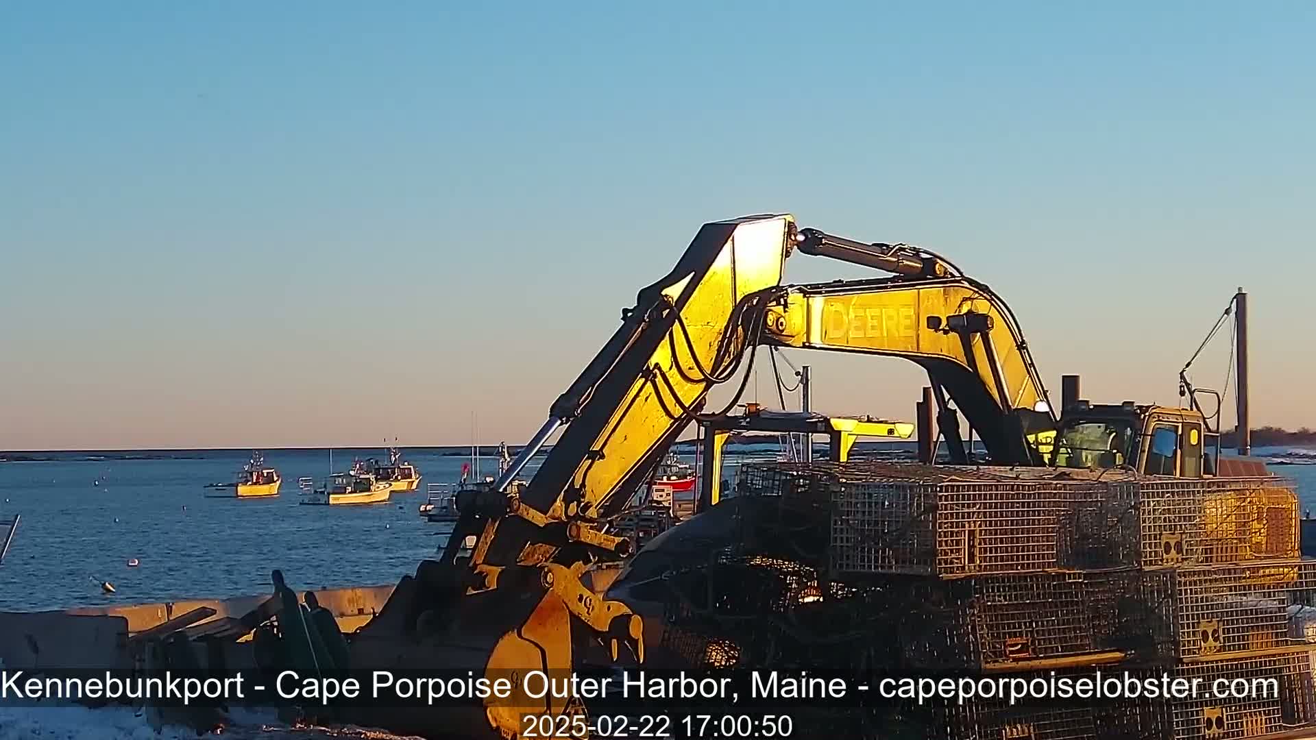 A yellow excavator sits on a dock at sunset, near several small fishing boats in calm water.