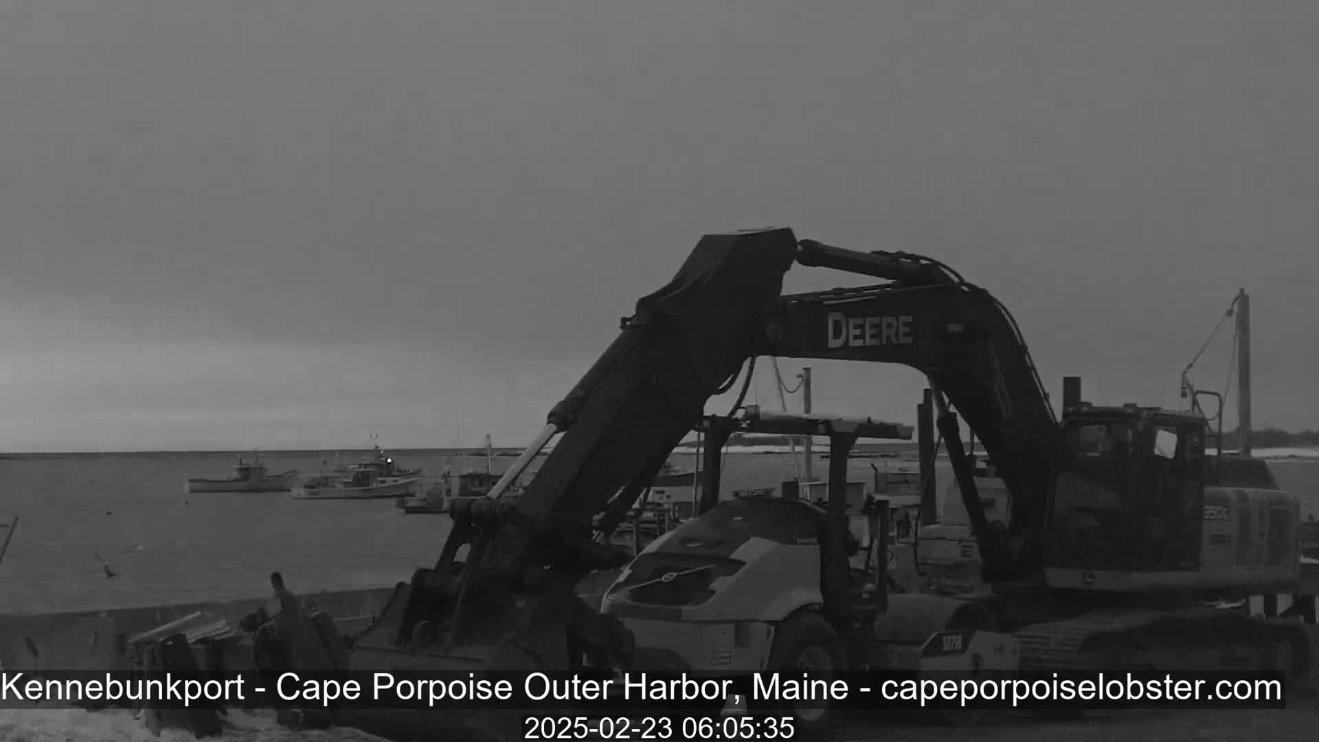 A large excavator sits in a harbor next to several small fishing boats under an overcast sky.