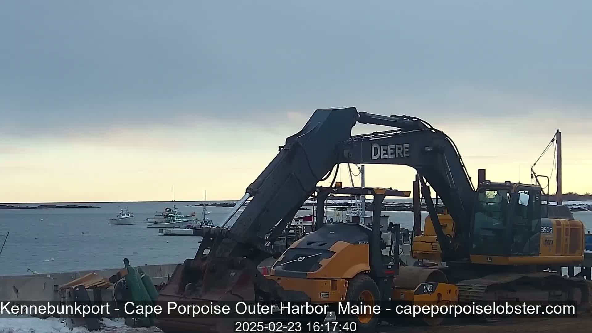 A large excavator and a smaller road roller are parked near a harbor with several boats, under a mostly cloudy, dusky sky.