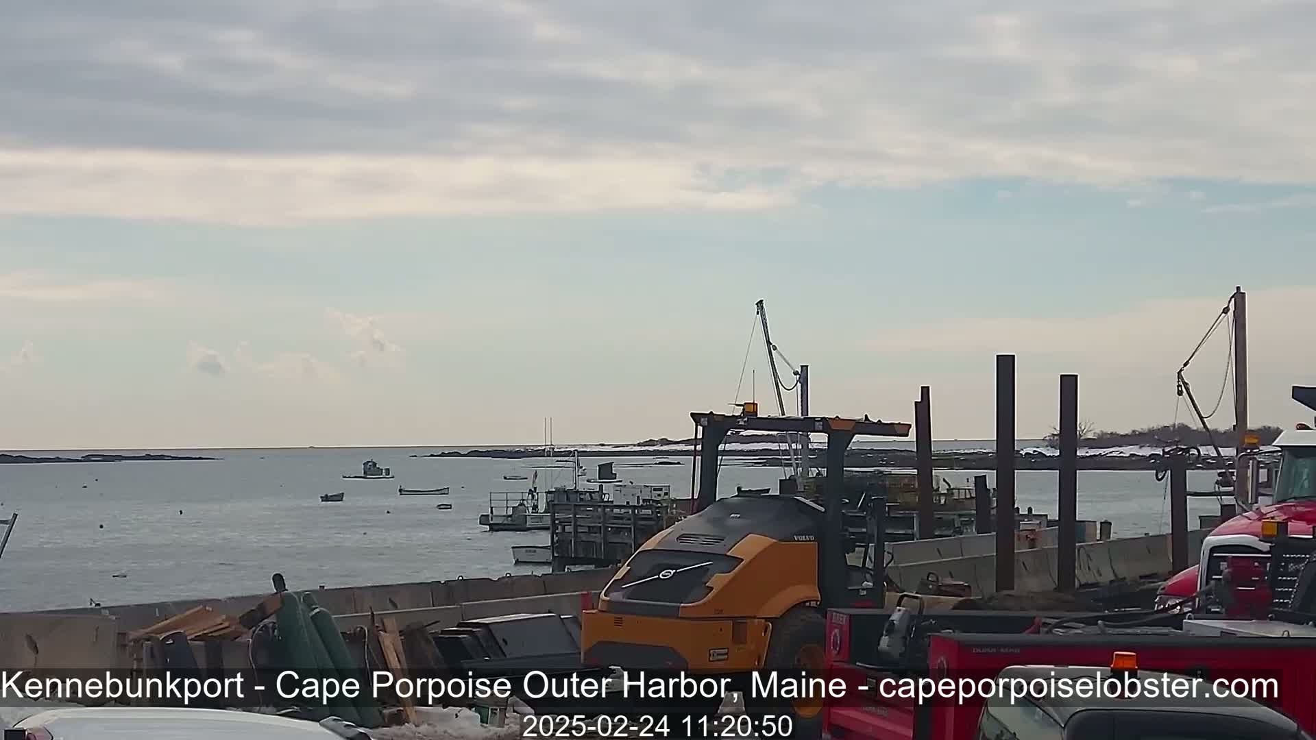 A mostly cloudy day at a harbor shows several small boats on the water, a yellow forklift, and other construction equipment near a pier.