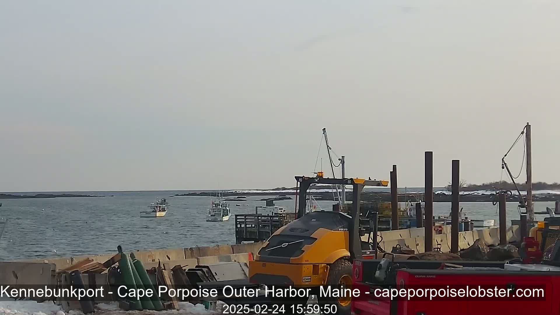 A calm, overcast day reveals several fishing boats in a harbor, with a yellow forklift and other equipment near a dock.
