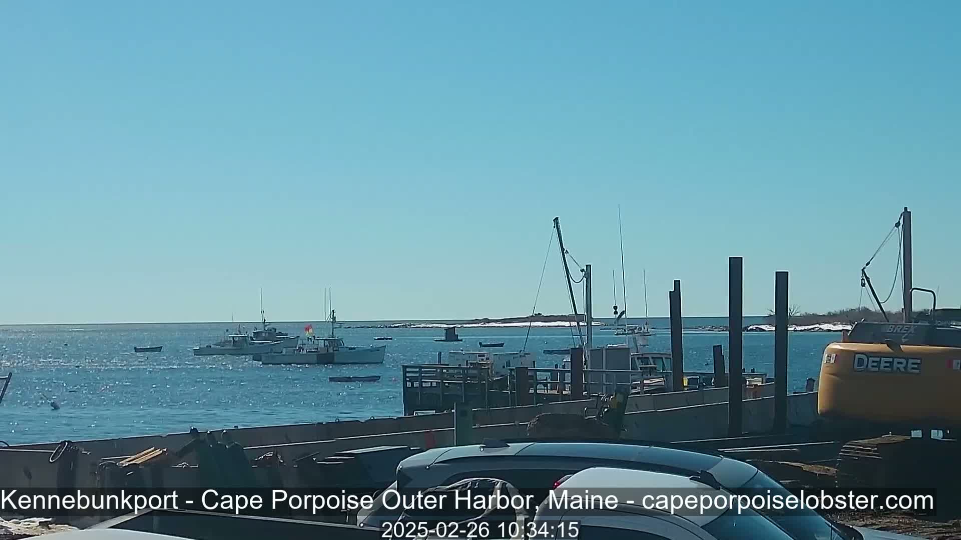 Several fishing boats are in a harbor on a sunny day, with a yellow excavator parked nearby.