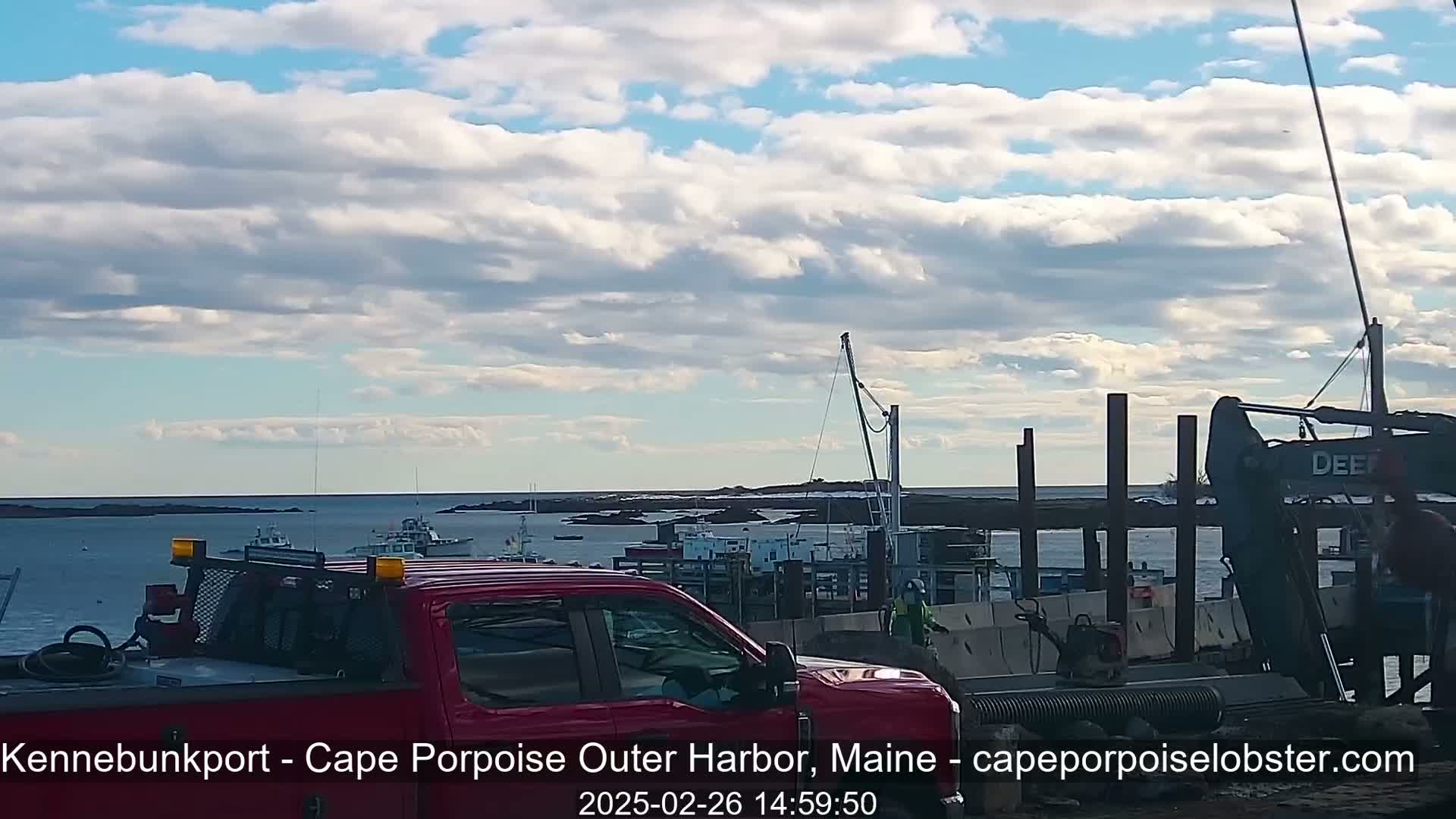 A red pickup truck is parked near a harbor where construction is taking place under a partly cloudy sky.