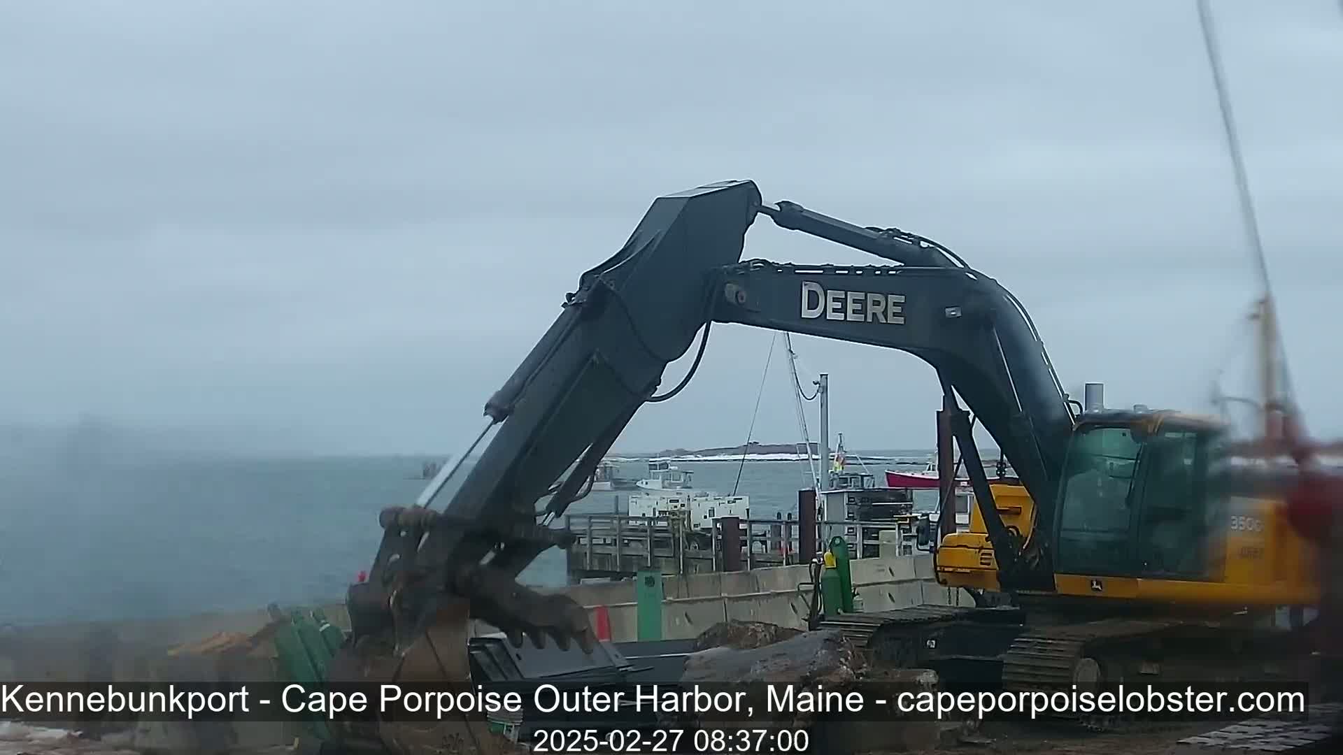 A large excavator is working near a harbor on an overcast day with several boats in the background.