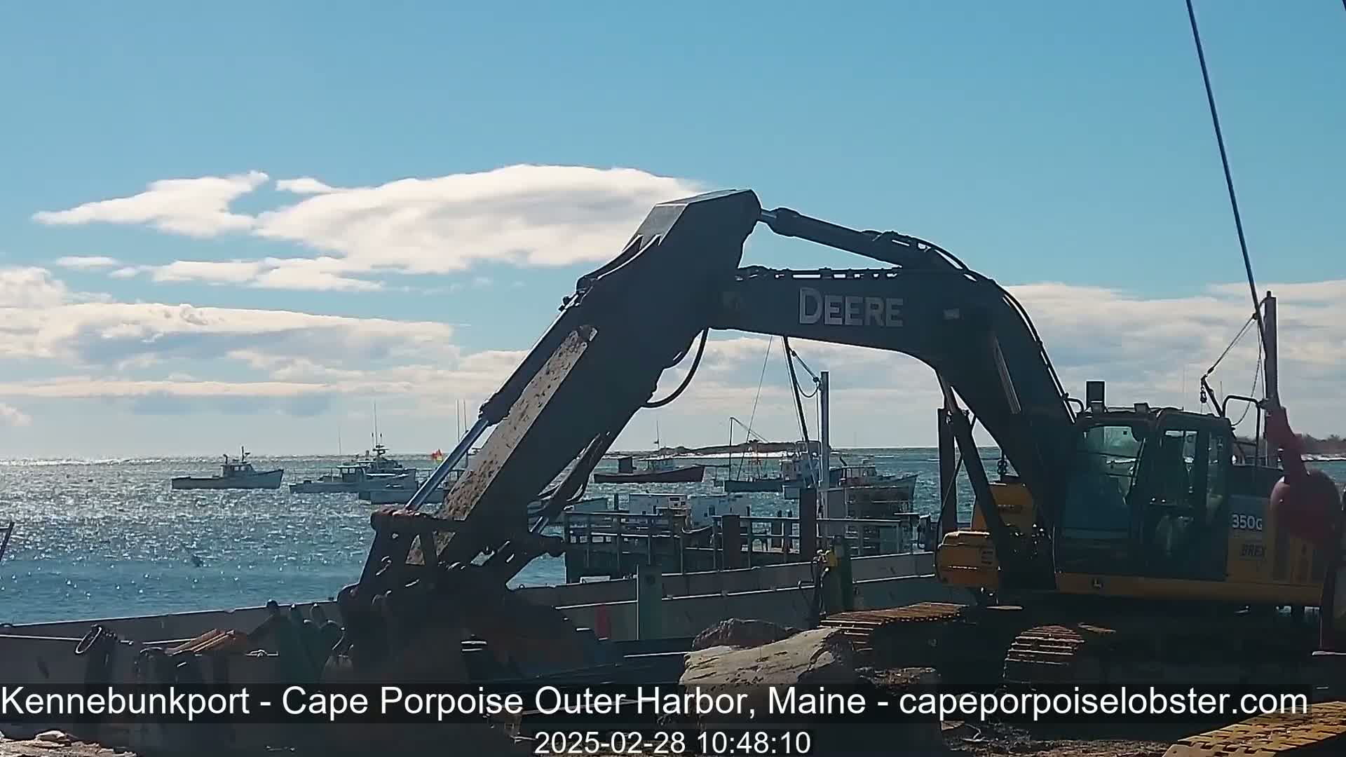 A large excavator sits on a barge in a harbor with several fishing boats in the background under a partly cloudy sky.