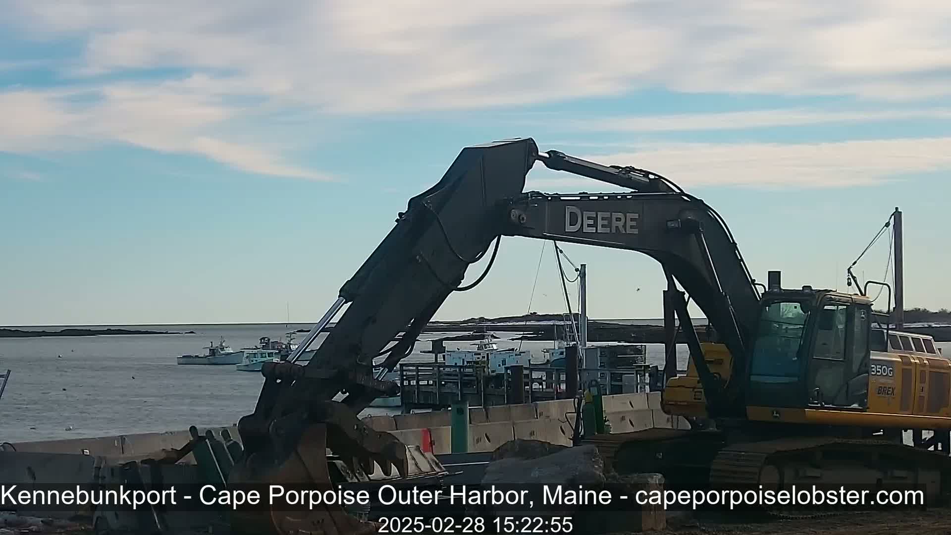 A large excavator sits near a harbor on a partly sunny day, with several fishing boats visible in the background.