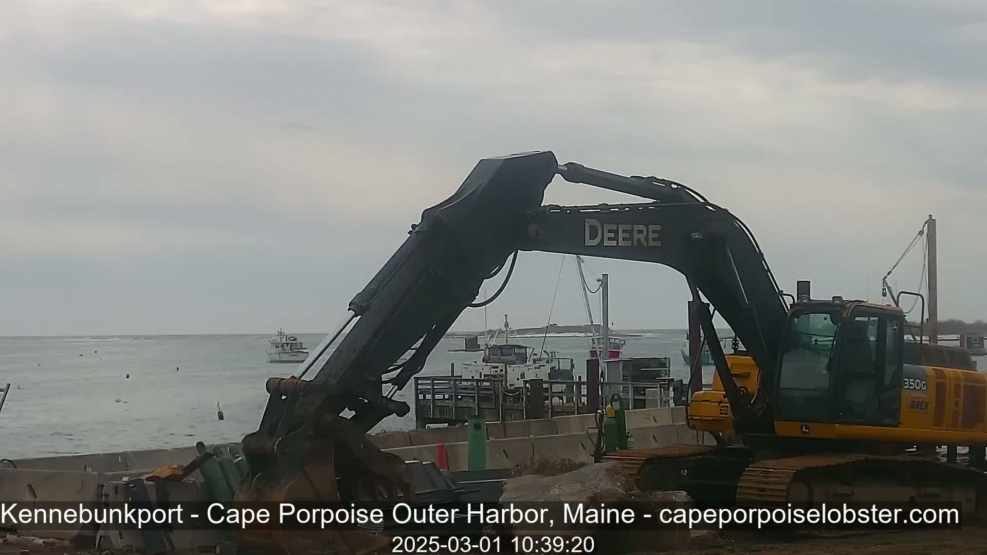 A large excavator is positioned near a harbor, with several boats visible in the overcast water.