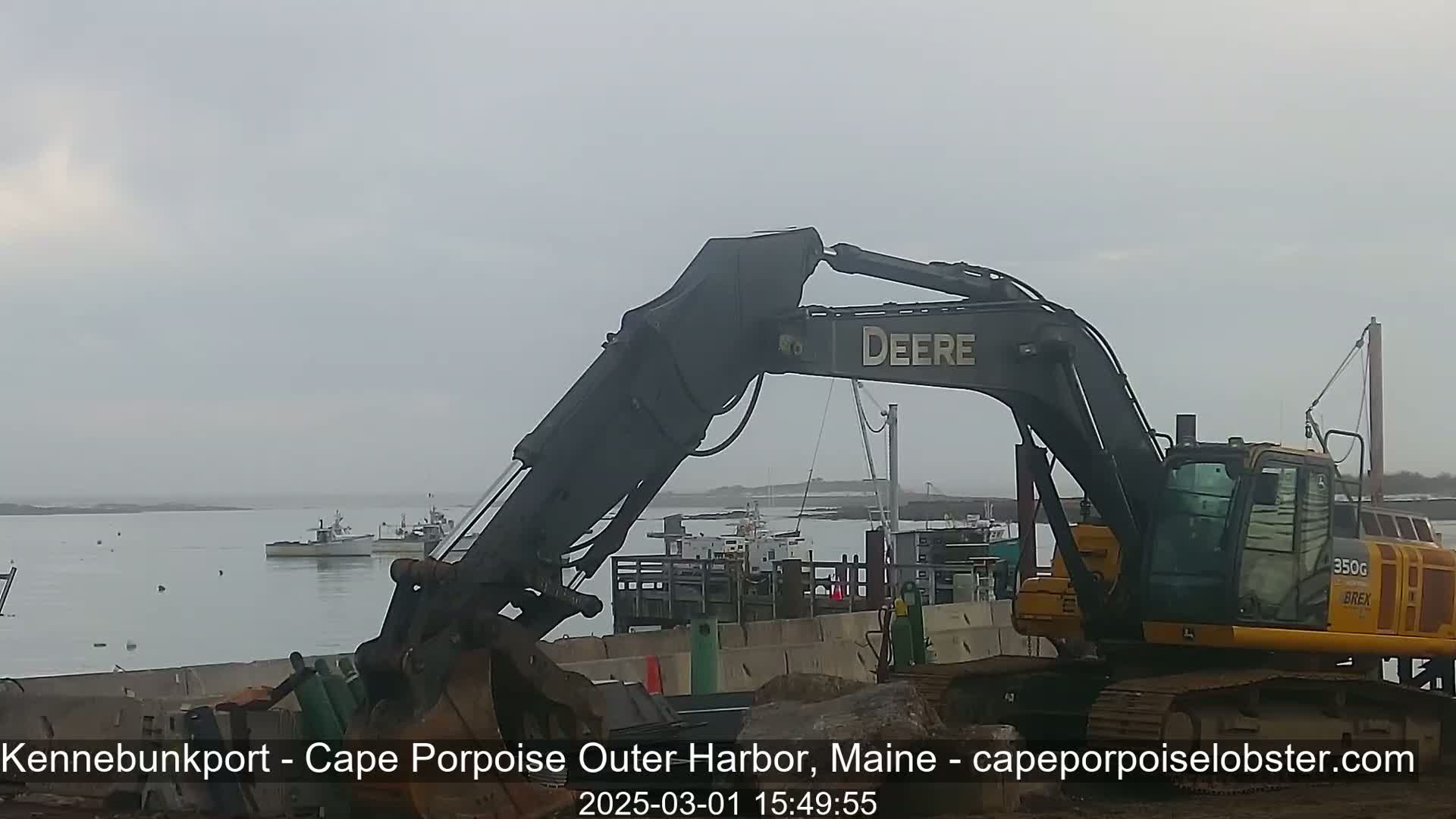 A large excavator sits near a harbor on an overcast day, with several boats visible in the water.