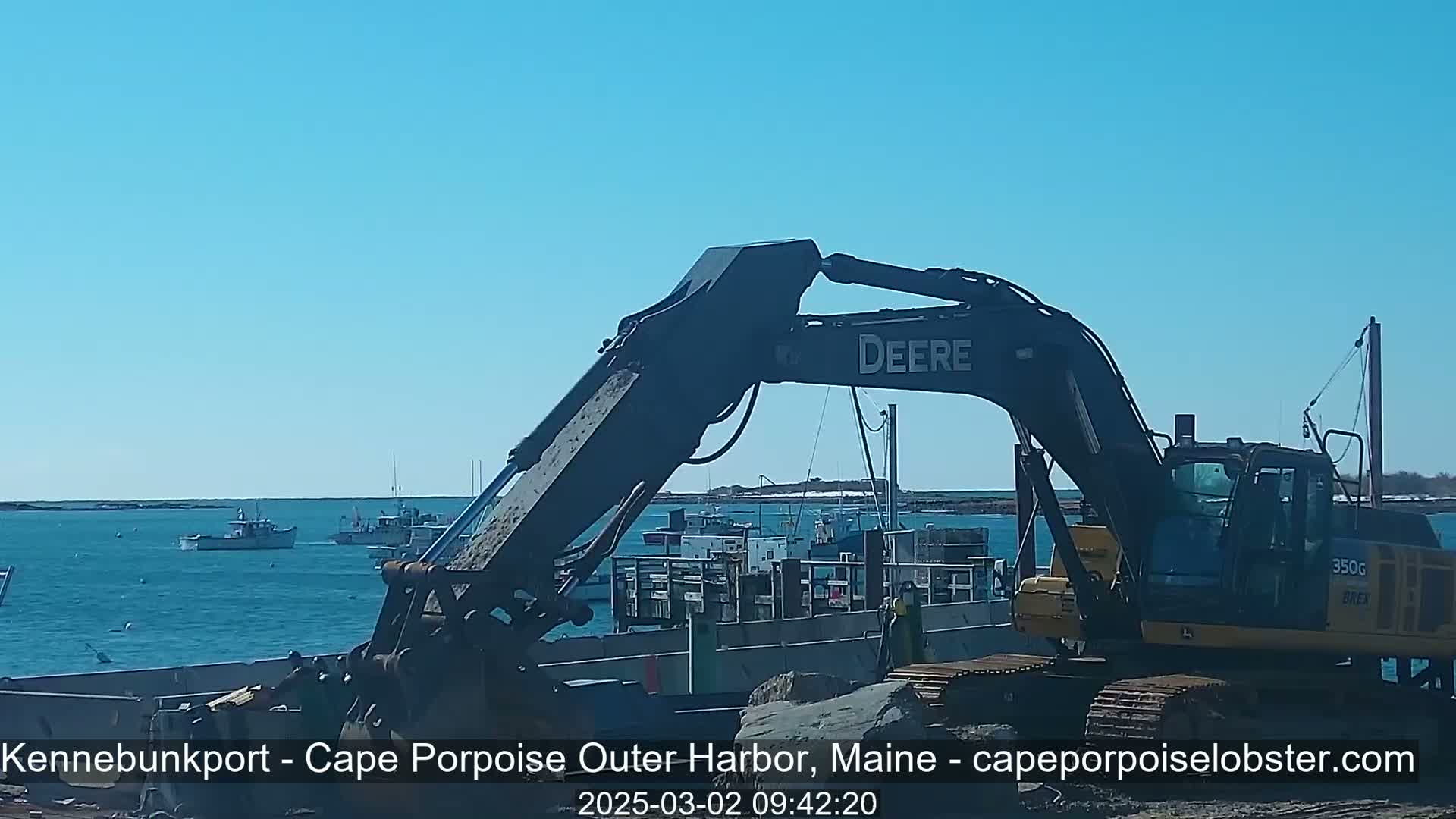 A large excavator sits on a barge in a harbor on a sunny day, with several boats visible in the background.