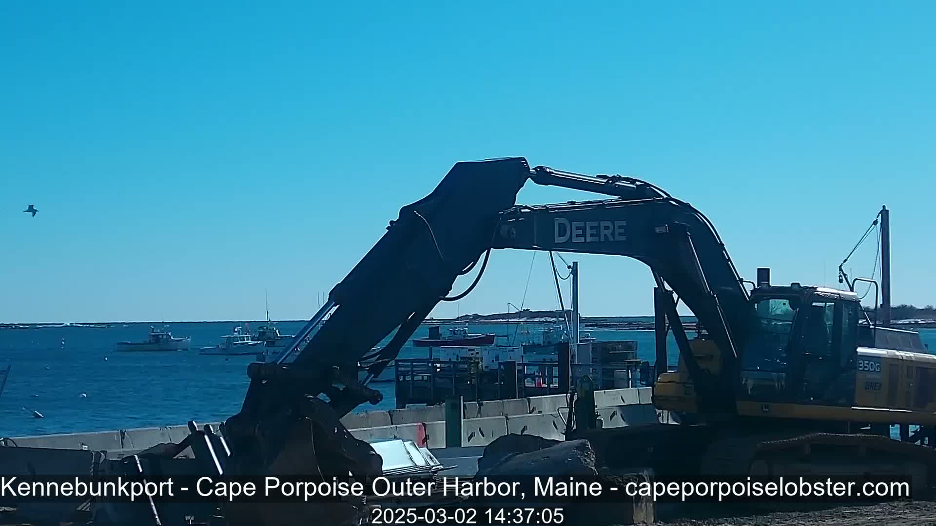 A large excavator sits near a harbor on a sunny day, with several boats visible in the calm water.