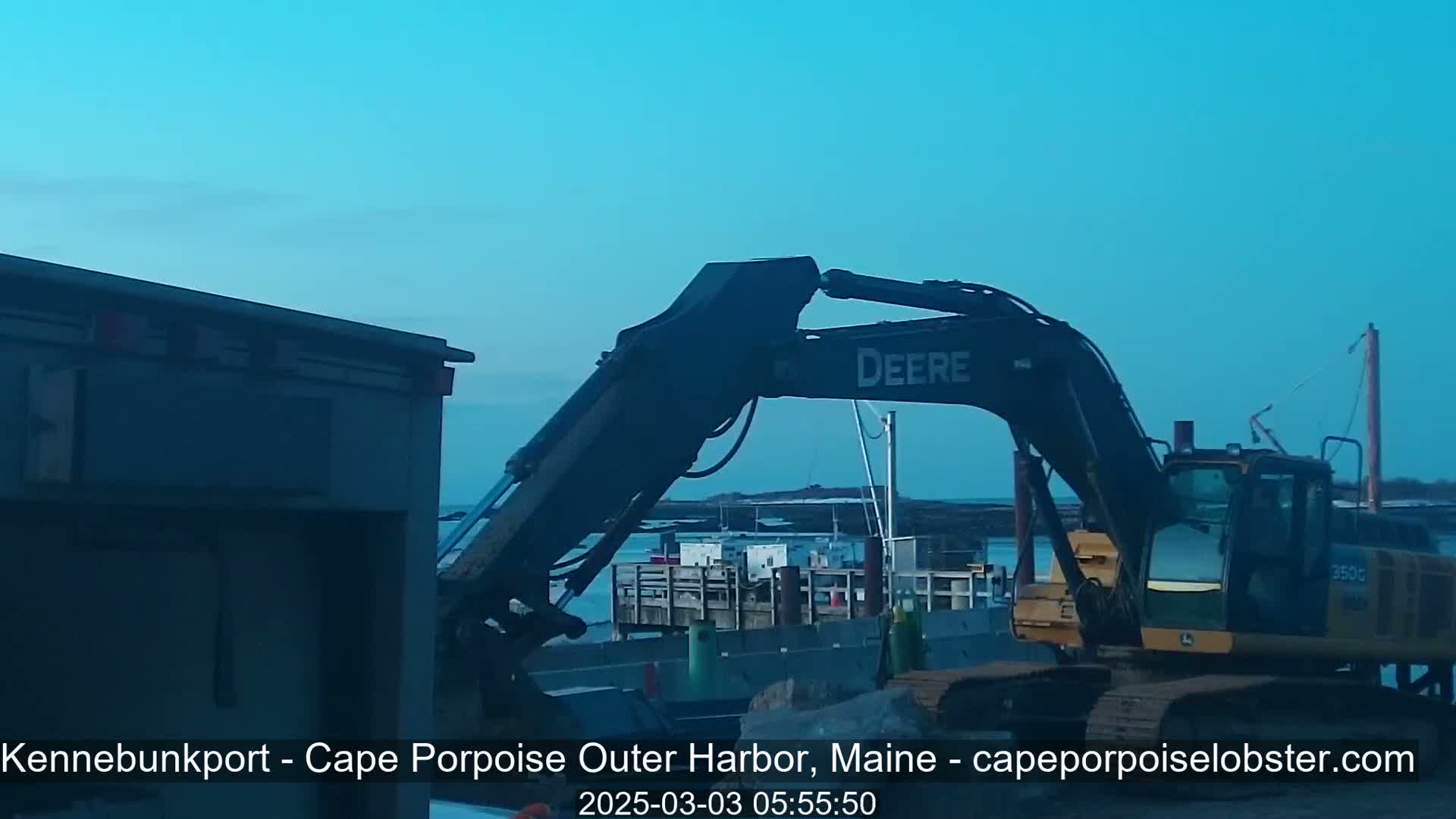 A large excavator sits near a body of water under a clear, light-blue sky.