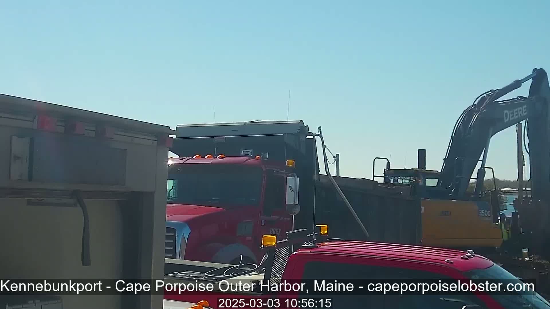 Under a clear blue sky, several large trucks and a construction excavator are parked near a body of water.