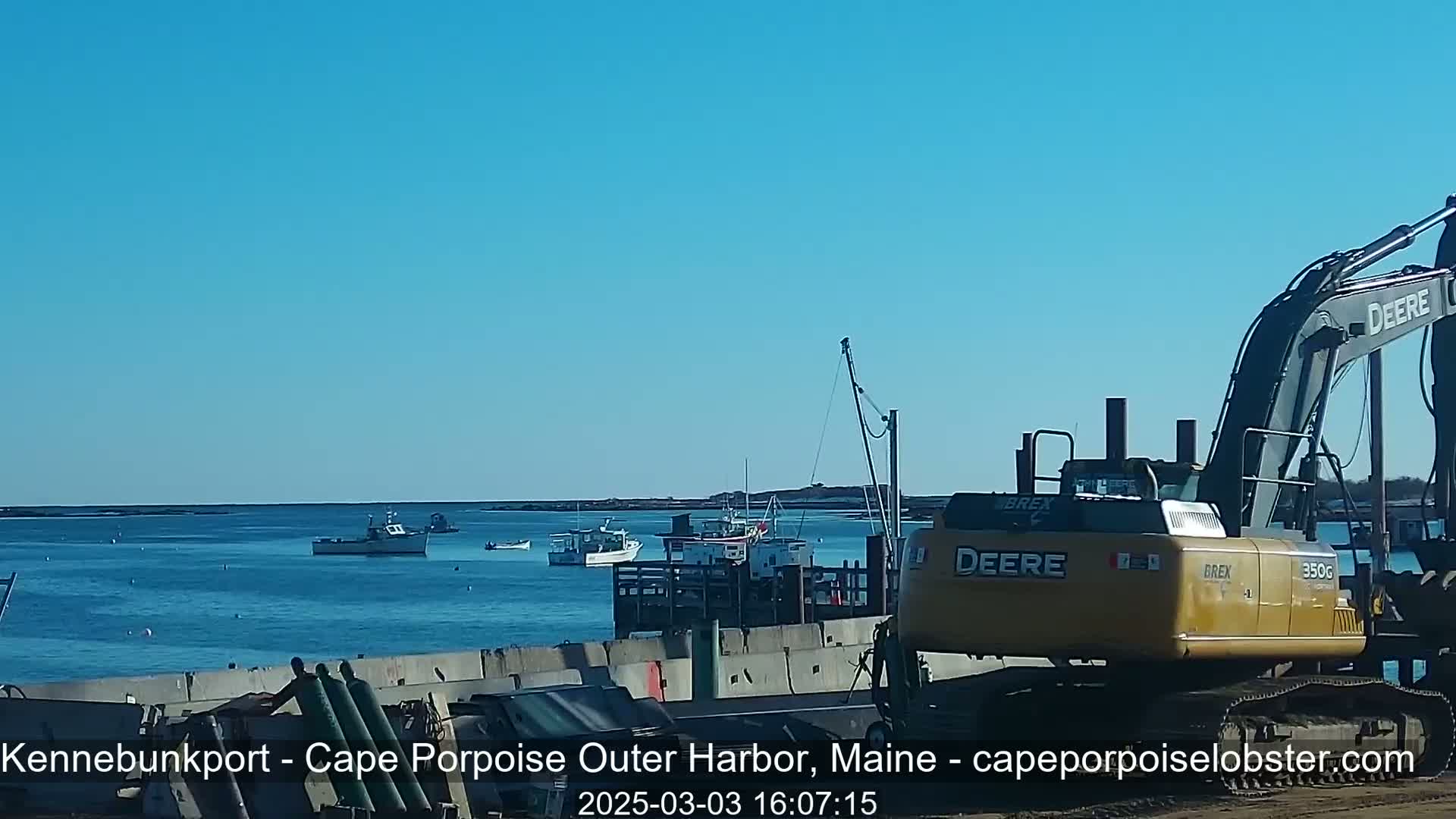 A yellow excavator sits near a harbor with several fishing boats on a clear, sunny day.