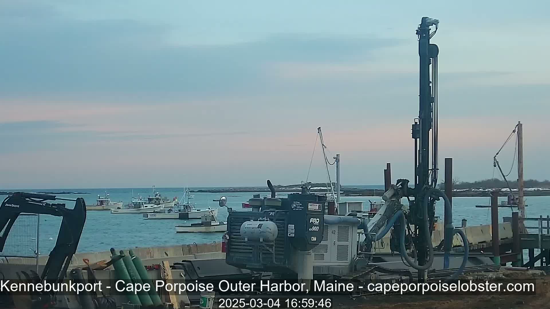 Several fishing boats are moored in a calm harbor at dusk, with construction equipment, including a tall drilling rig, visible on the shore.