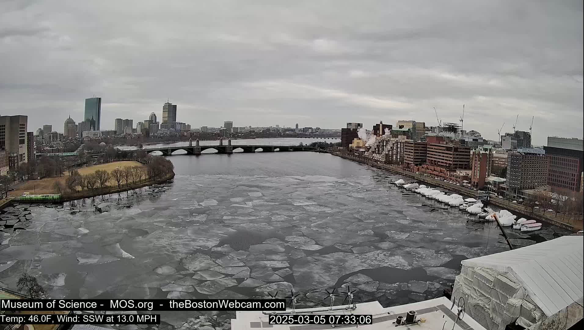 A partially frozen river flows past a city skyline under an overcast sky.