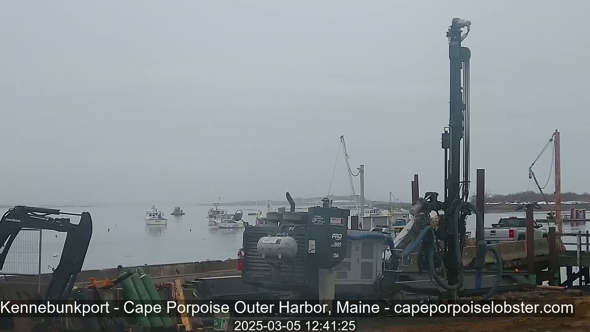 A large drilling rig sits on a dock overlooking a calm, foggy harbor with several small fishing boats.
