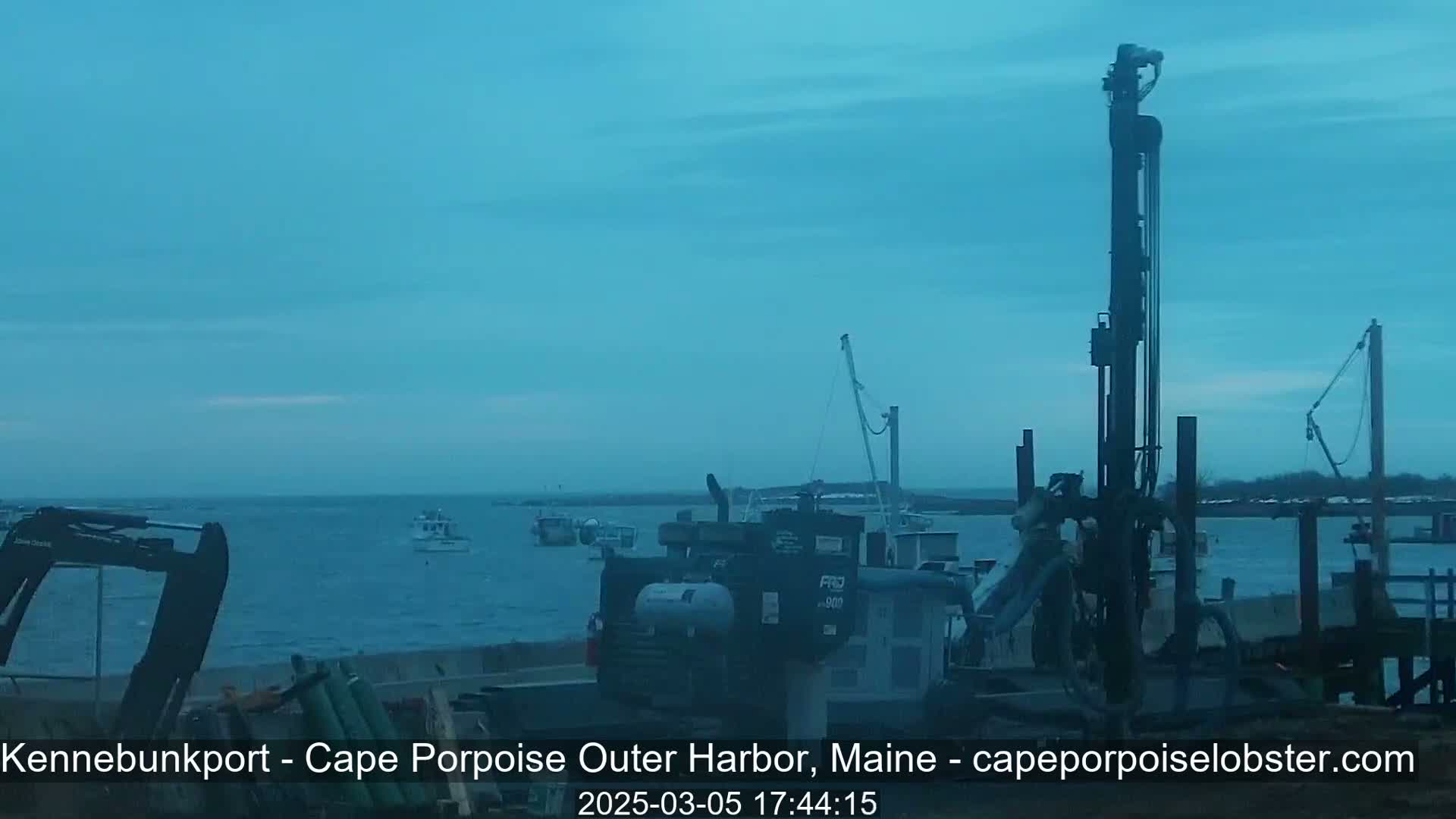 Several boats are in a harbor under a dusky blue sky, with construction equipment visible on the shore.