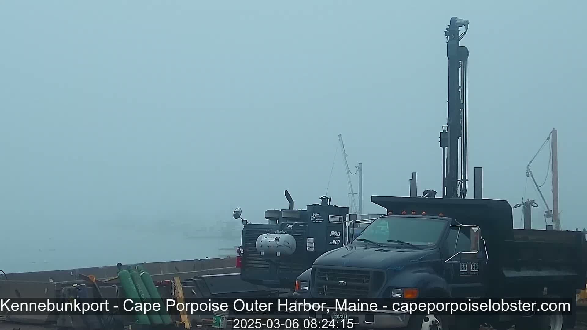 A foggy harbor scene features a dark-colored dump truck with a tall drilling rig in the background, along with other construction equipment and materials.