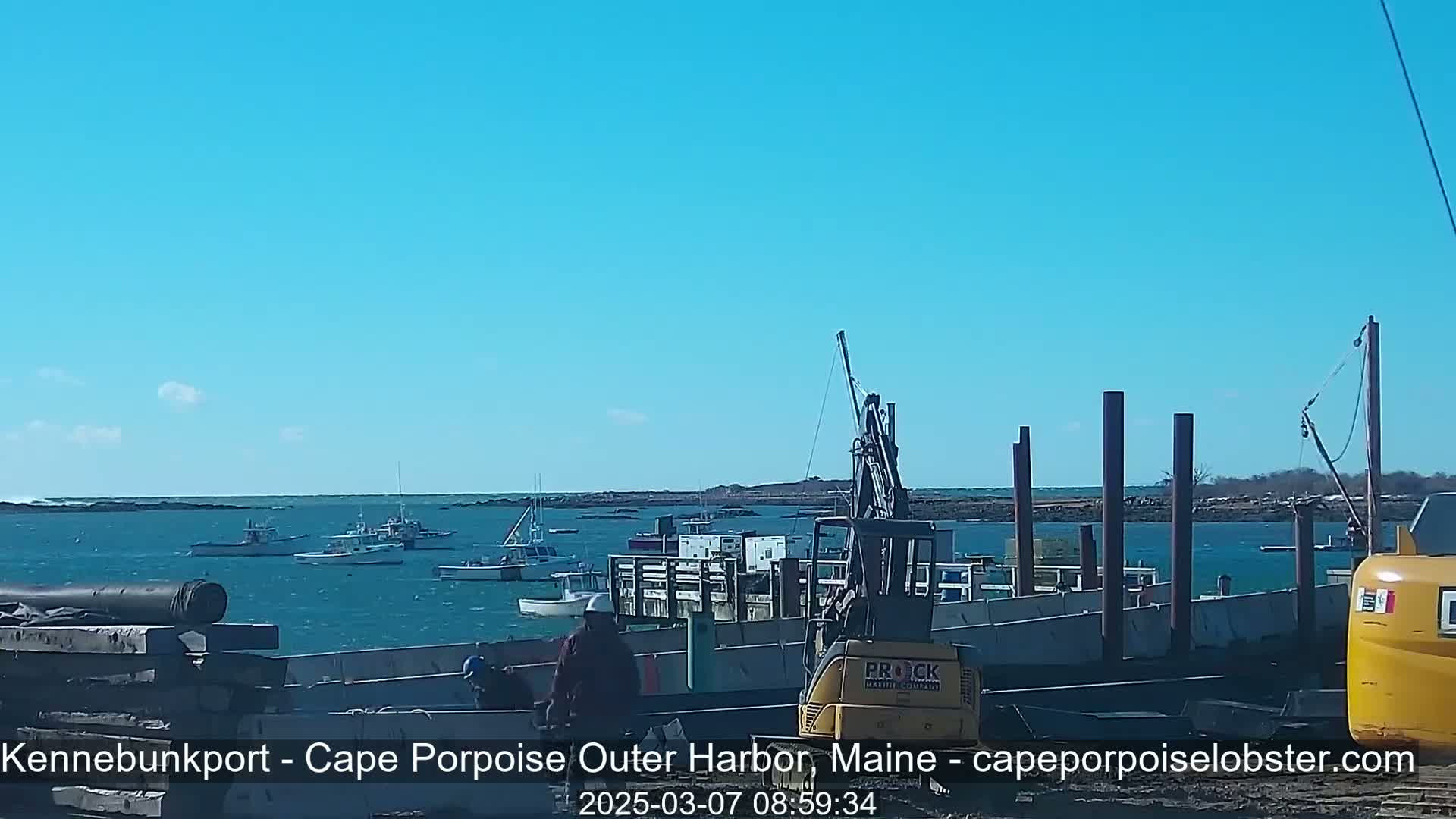 Under a clear blue sky, several fishing boats are moored in a harbor near a construction site where a yellow forklift and other heavy machinery are present.