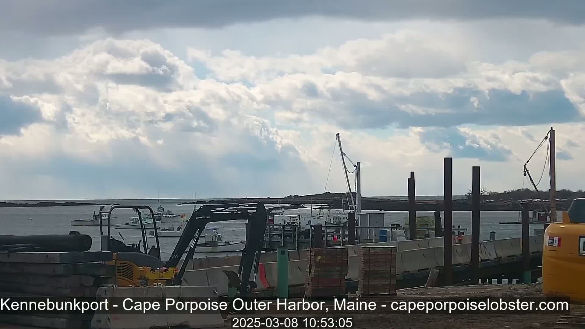 A partly cloudy day shows a harbor with several boats, construction equipment, and partially built docks.