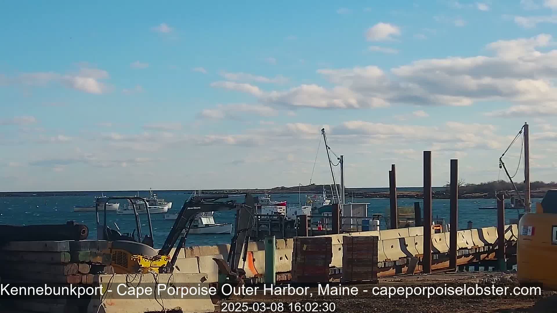 A partly cloudy day shows several boats in a harbor with construction equipment and materials near a pier.