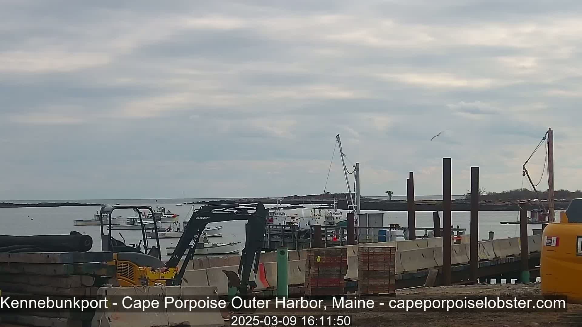 Several boats are moored in a harbor under an overcast sky, with construction equipment and materials visible on a nearby dock.