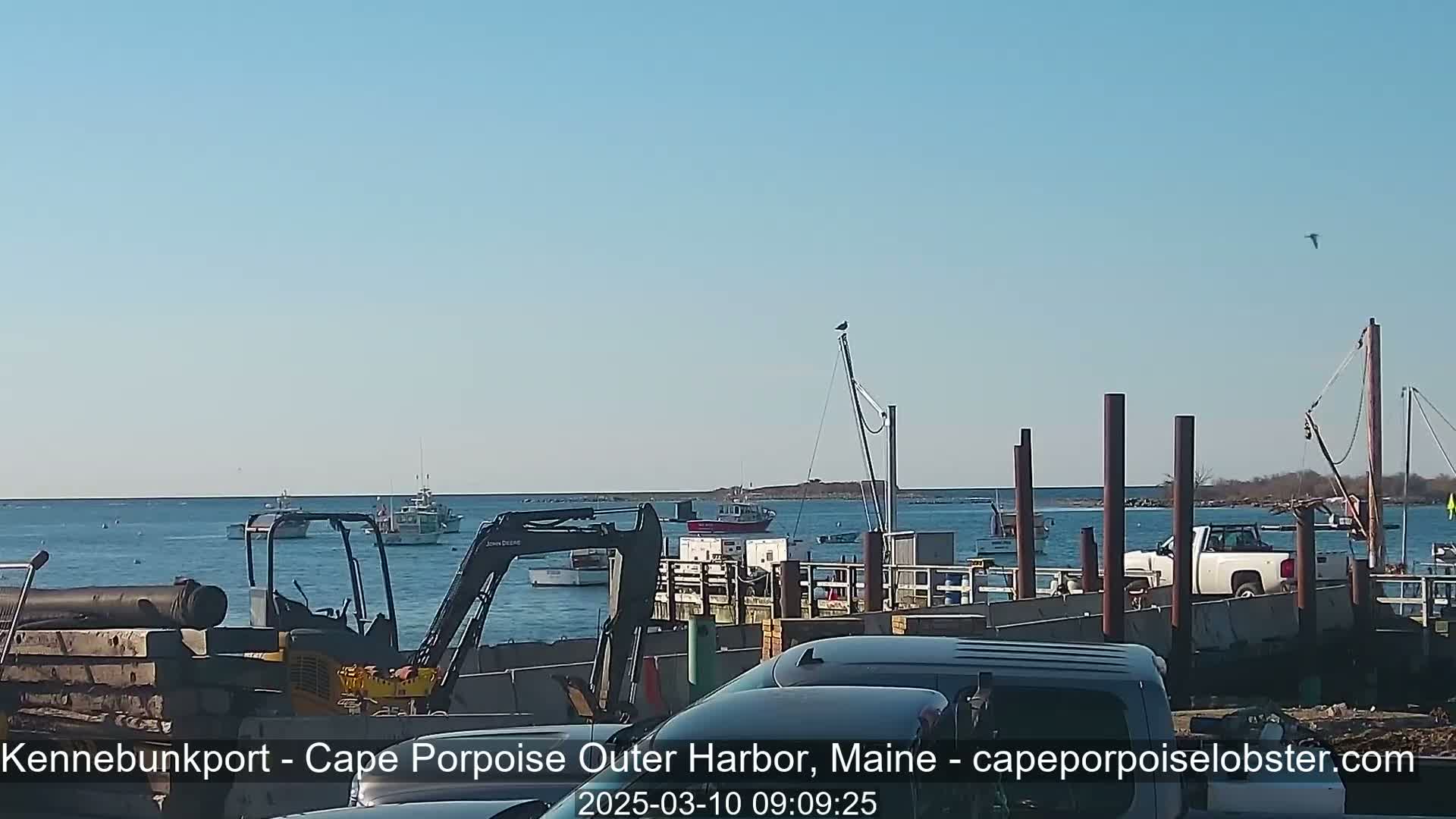 A harbor scene on a sunny day shows several boats in the water, construction equipment and vehicles on a dock, and a pickup truck parked nearby.