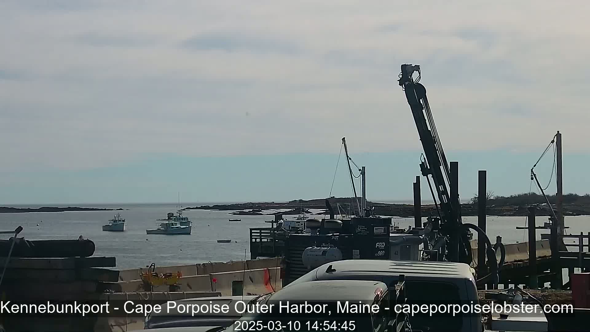 A calm, sunny day shows several fishing boats in a harbor, with construction equipment and vehicles on a dock in the foreground.