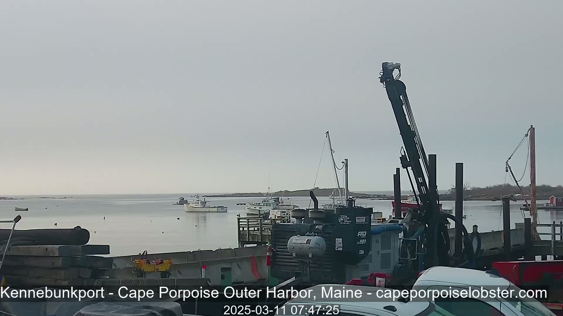 Several boats are moored in a calm harbor under an overcast sky, with a large piece of industrial equipment visible in the foreground.