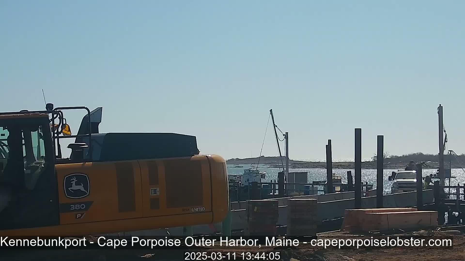 A yellow excavator is parked near a harbor under sunny skies, with a boat and construction materials visible in the background.