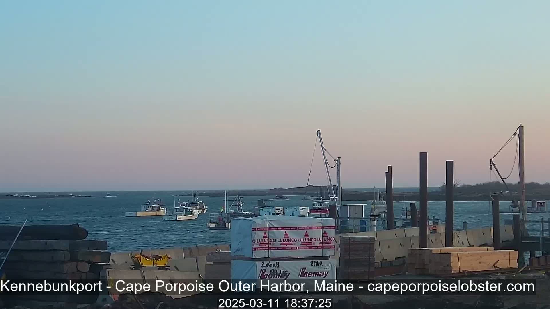 Several fishing boats are in the water near a harbor under a pale pink and blue twilight sky.