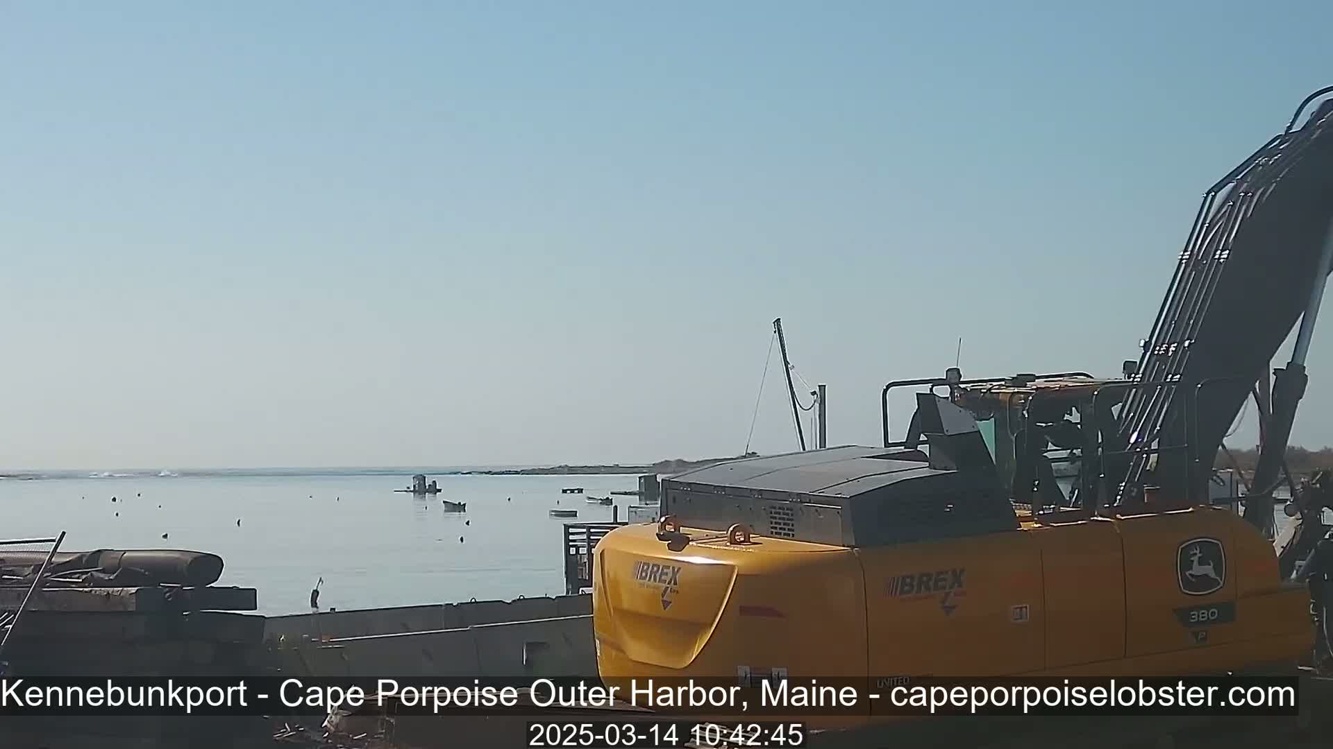 A yellow excavator is parked near a calm body of water on a sunny day, with several small boats visible in the distance.