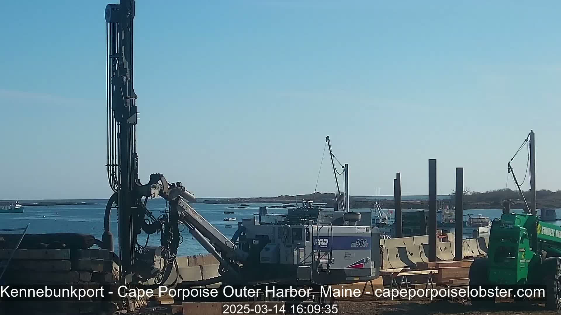 A large drilling machine is positioned near a body of water under a clear blue sky, with other construction equipment and boats visible in the background.