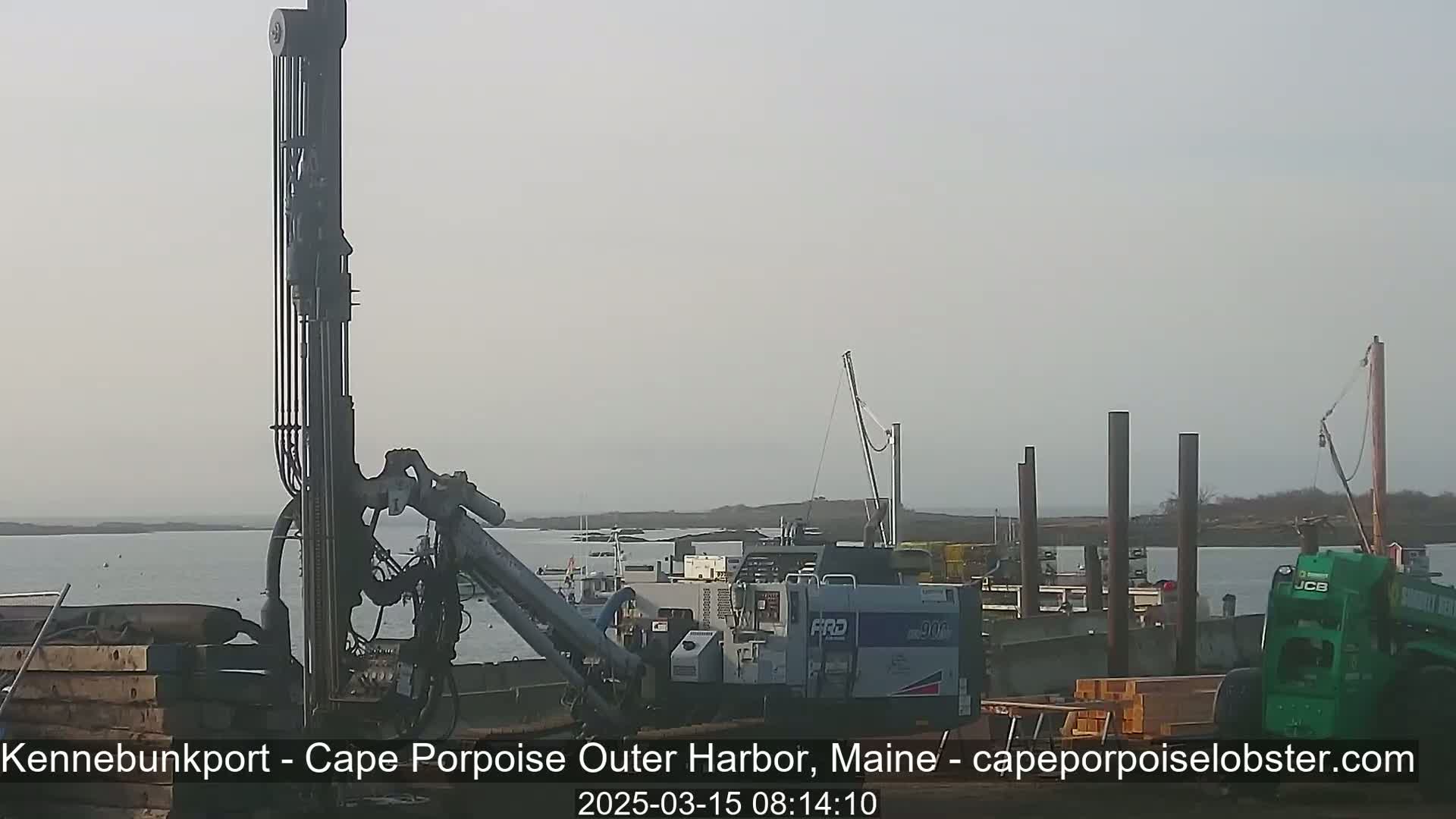 Construction equipment is positioned on a dock overlooking a calm body of water under an overcast sky.