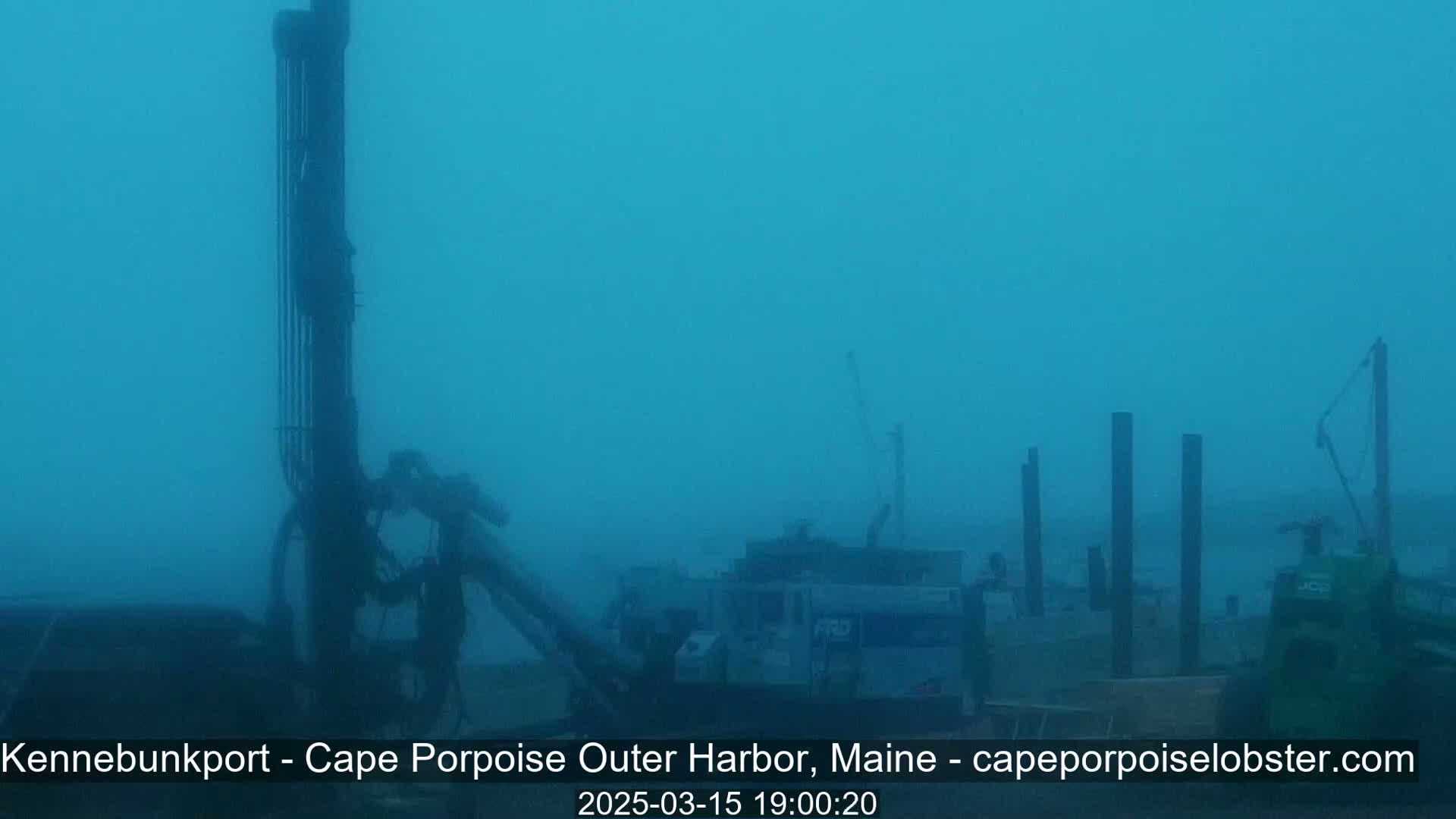 Heavy fog obscures several pieces of heavy machinery and tall poles near a body of water.