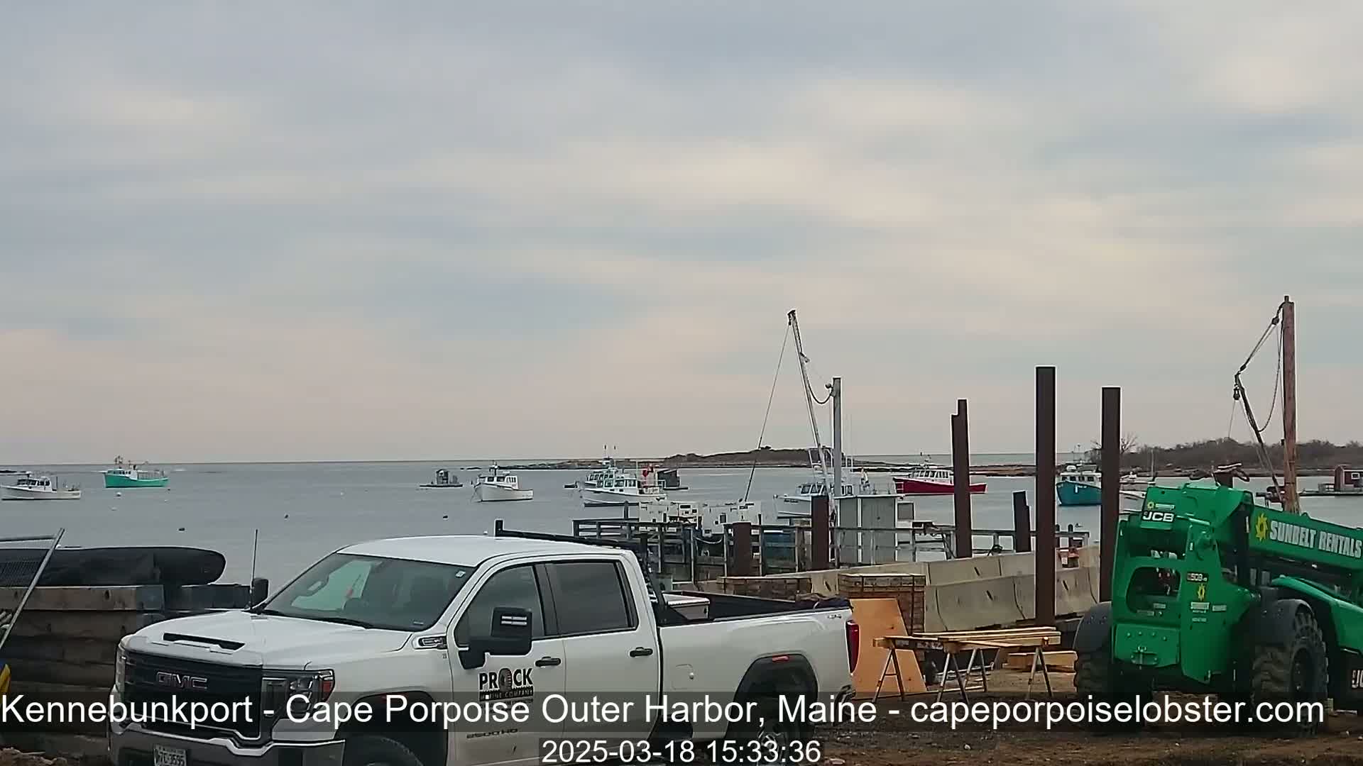 A harbor scene under an overcast sky shows several boats in the water, a white pickup truck, a green telehandler, and construction materials near a dock.