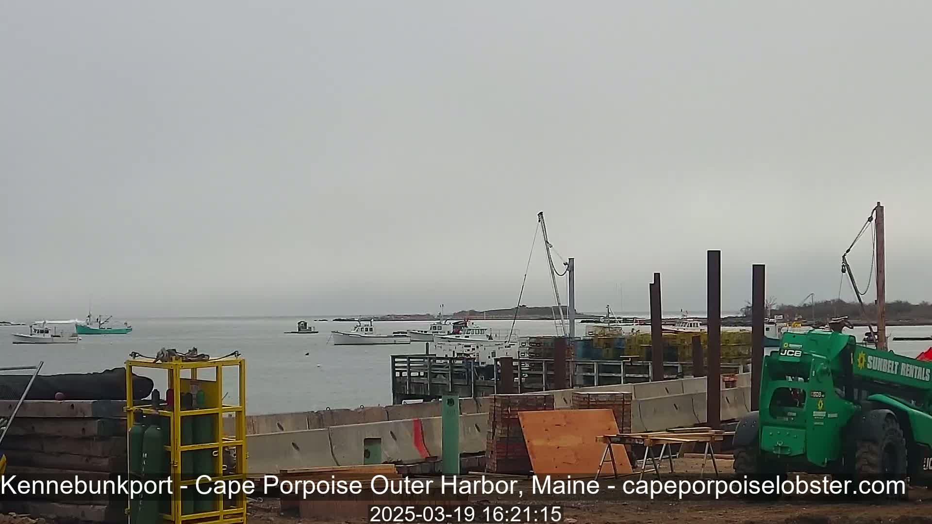 Several boats are moored near a harbor under an overcast sky, with construction equipment and materials visible on the shore.