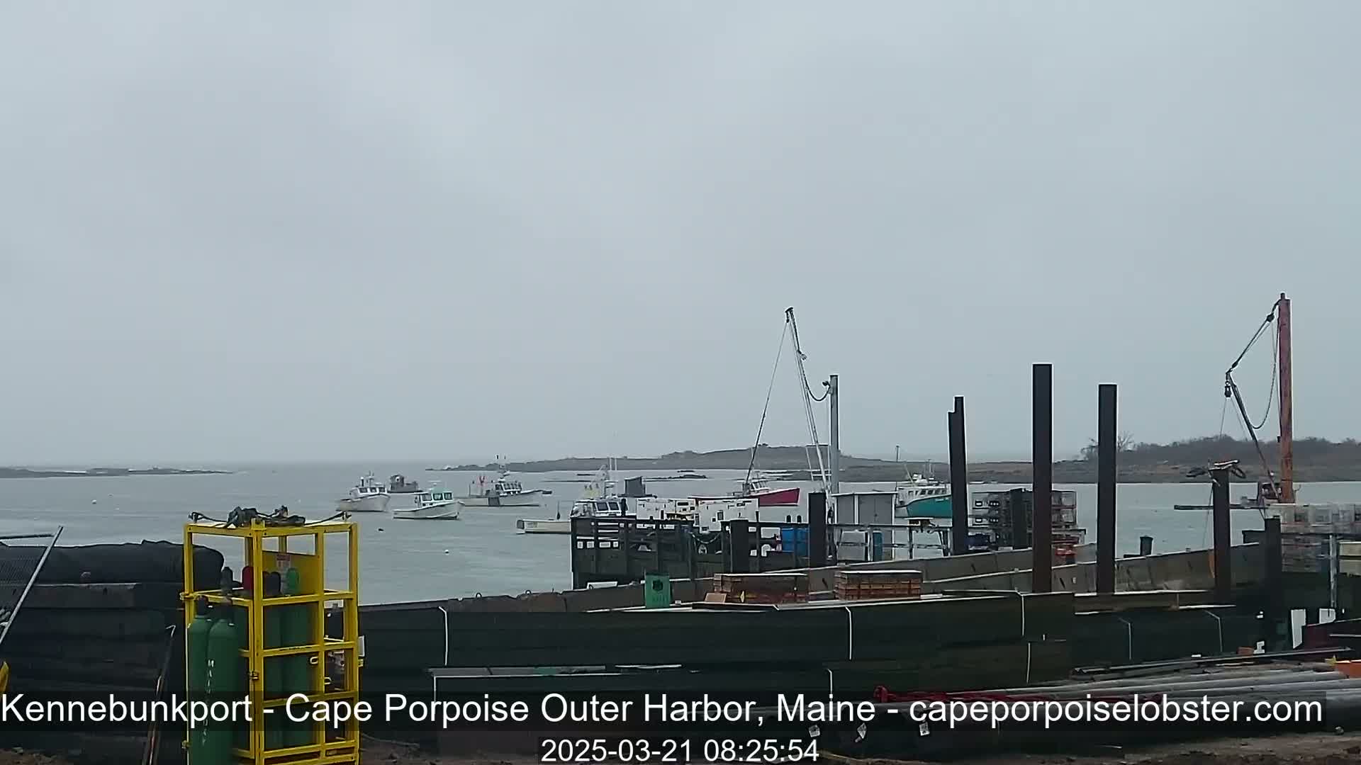 Several boats are moored in a harbor on an overcast day, with construction underway on a dock in the foreground.