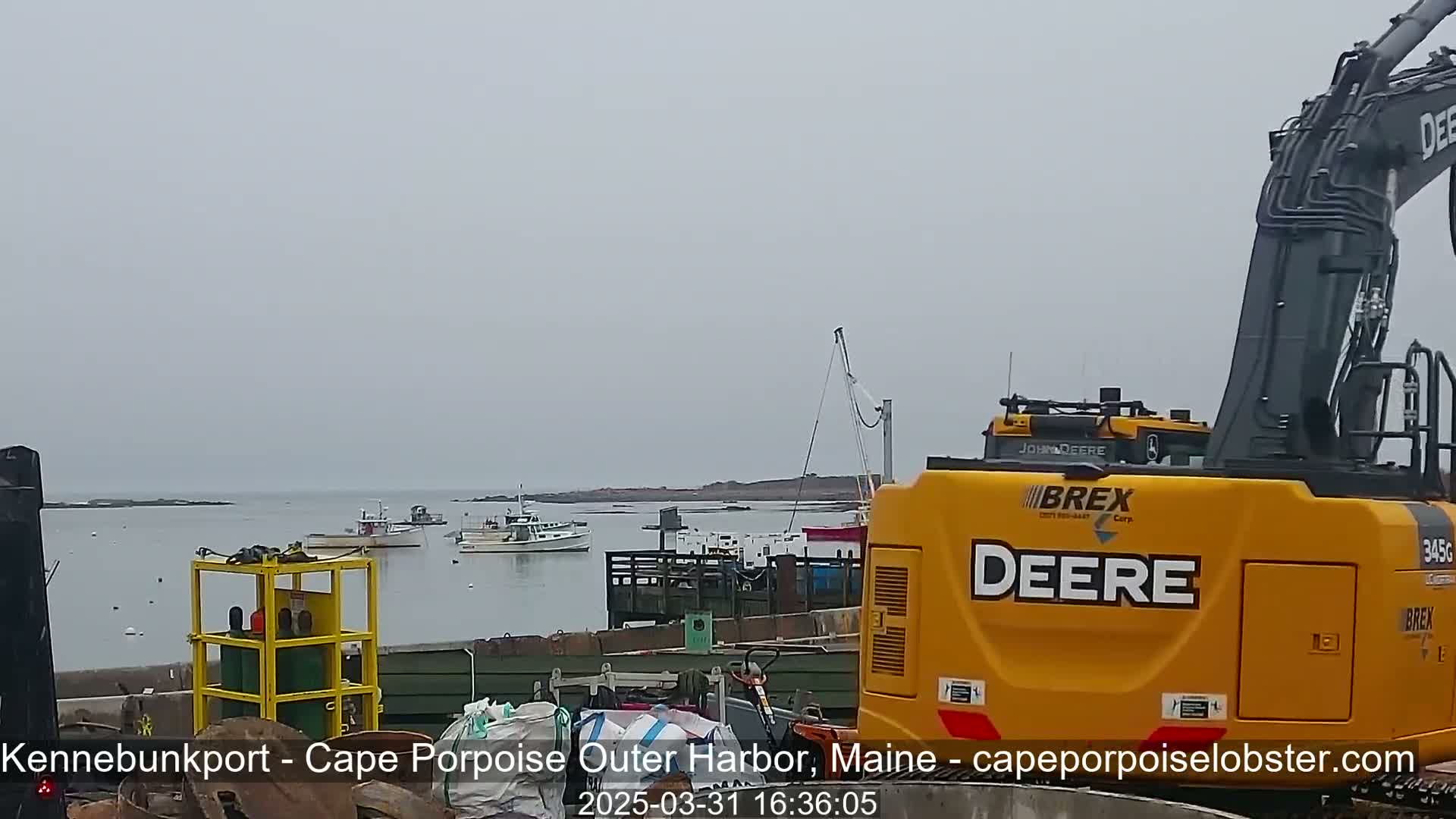 A yellow excavator sits in the foreground overlooking a calm harbor with several small fishing boats and a pier under an overcast sky.