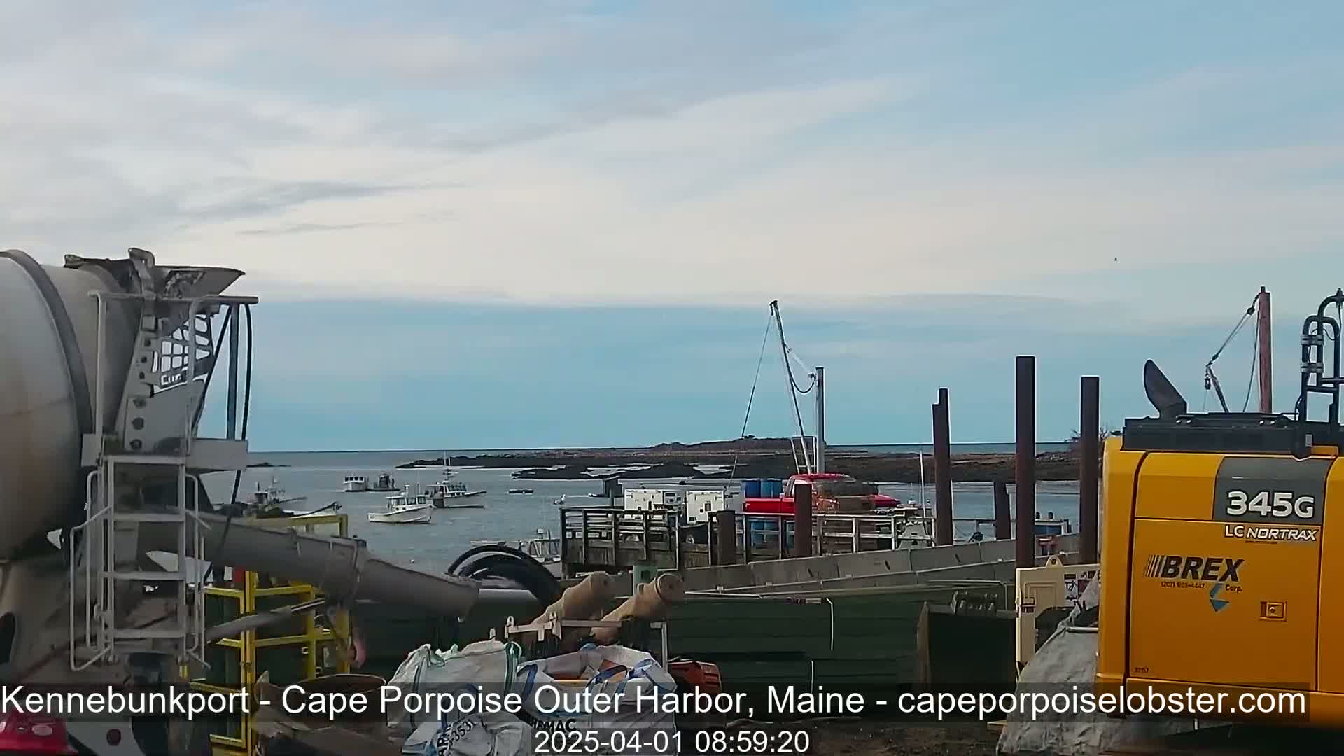 A cement mixer and excavator are positioned near a harbor, where several boats are moored under a mostly clear sky.