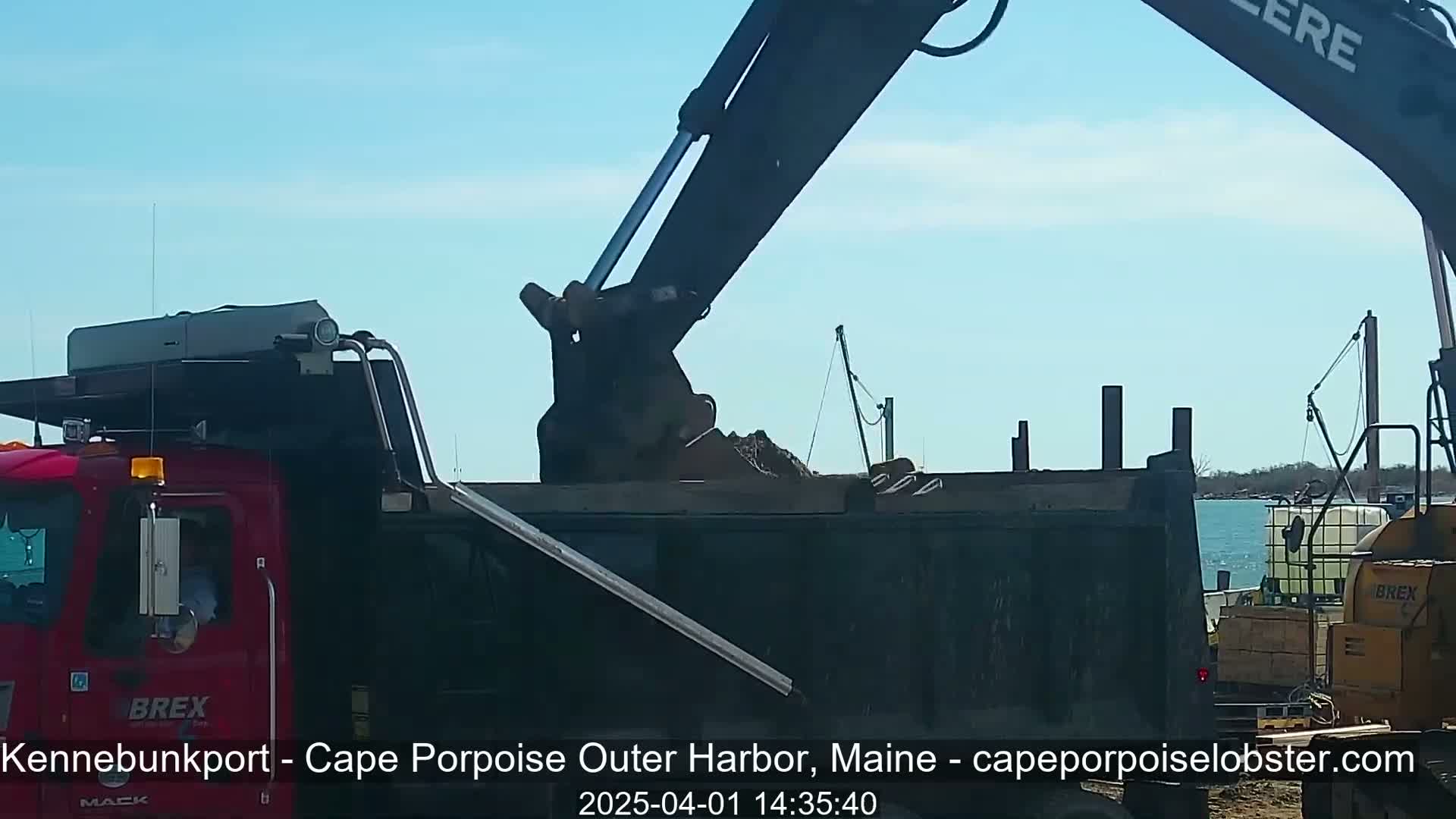 An excavator is dumping dirt into the back of a dump truck on a sunny day near a body of water.