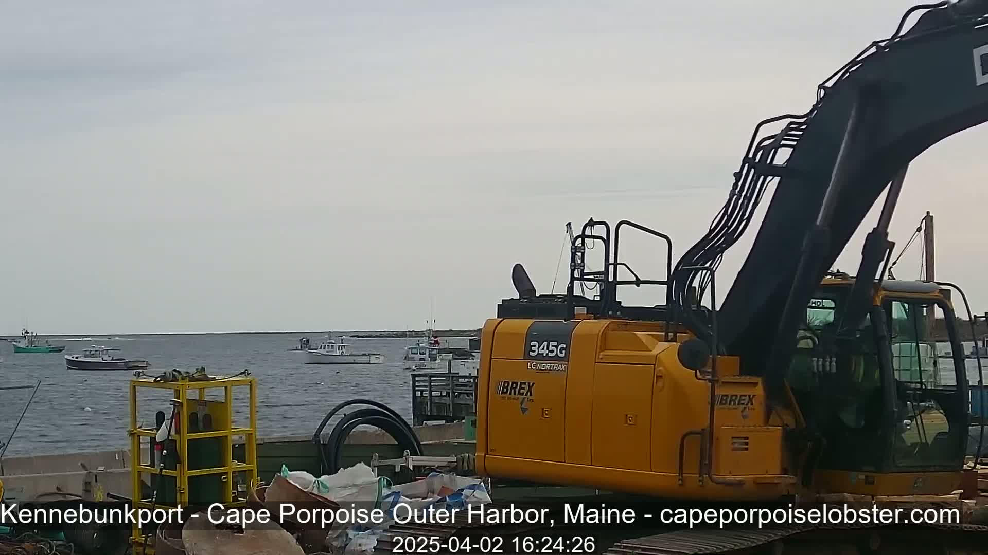 A yellow excavator sits on a dock overlooking a calm body of water with several boats, under an overcast sky.