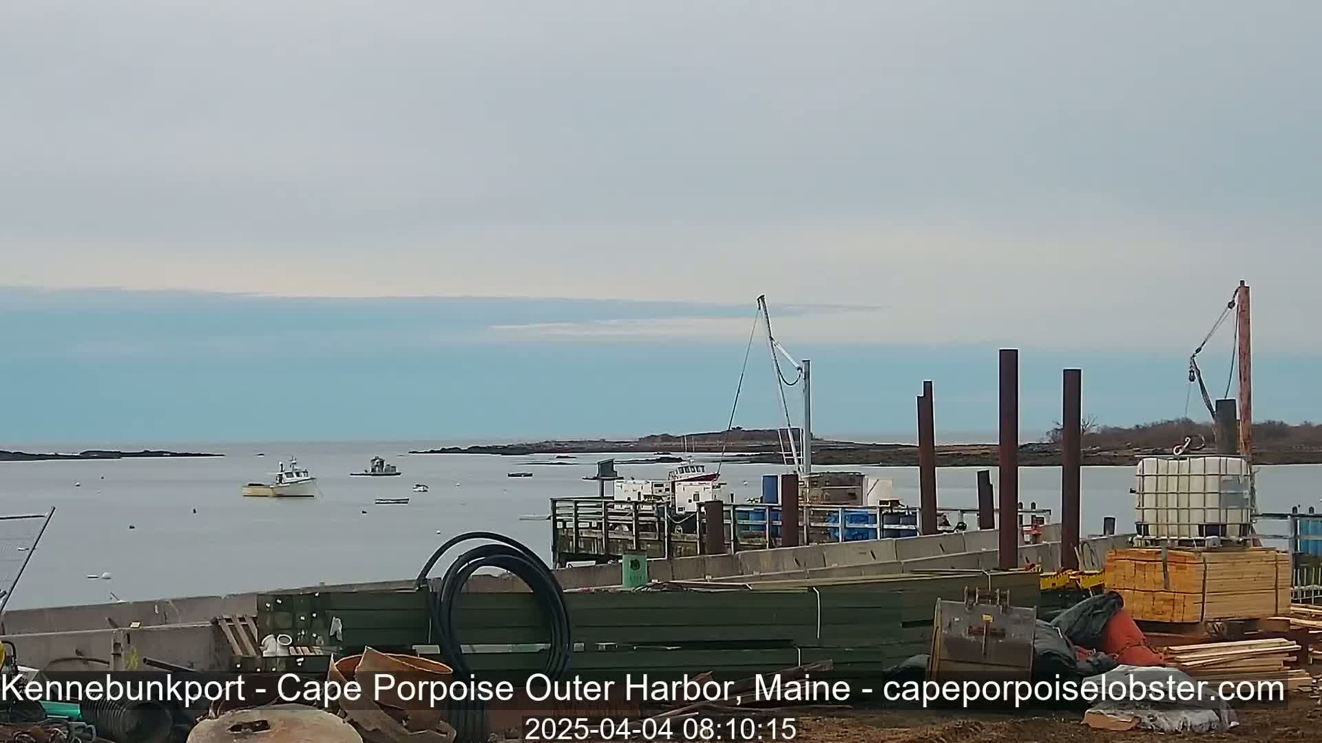 A calm, overcast day shows a harbor with a boat docked at a pier undergoing construction, another boat in the distance, and a small island in the background.
