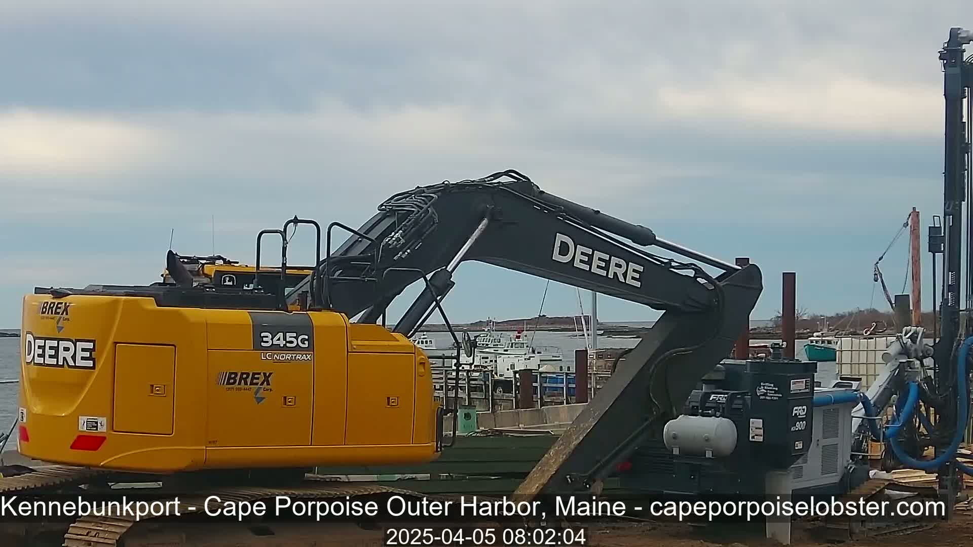A yellow Deere excavator and other heavy machinery are parked near a harbor on an overcast day.