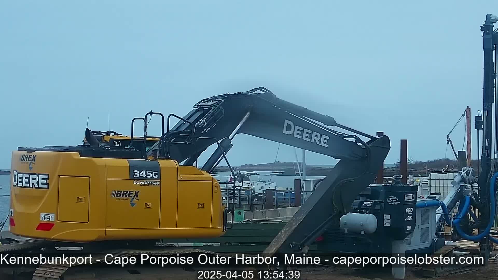 A yellow and gray Deere excavator and a drilling rig are parked near a harbor on an overcast day.