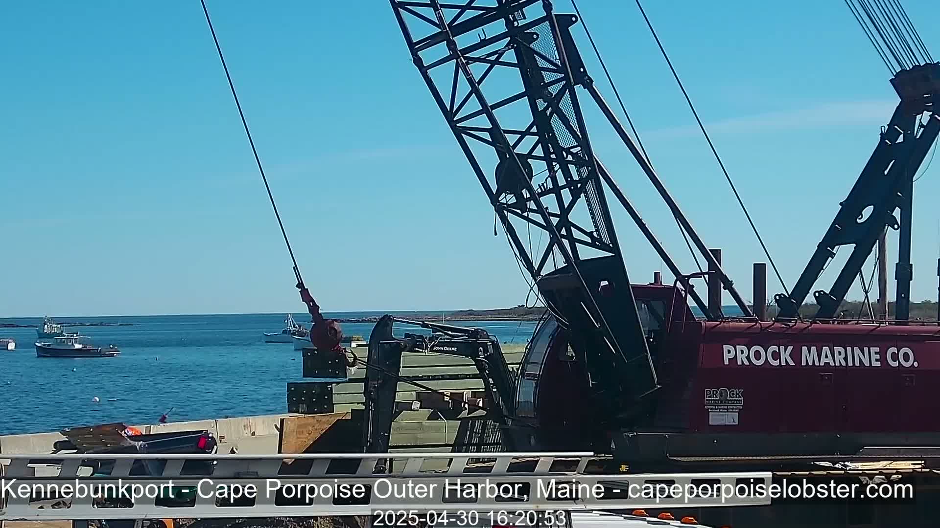 A large red crane lifts a stack of lumber near a harbor on a sunny day, with several small boats visible in the calm water.