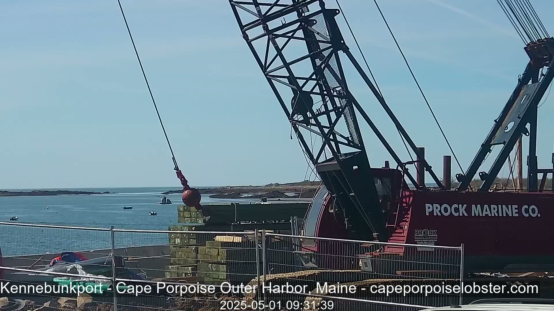 A large red crane on a construction site near a body of water lifts a heavy ball, under a clear blue sky.