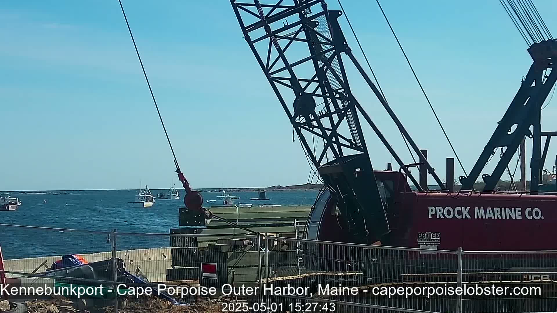 A large crane is positioned near a harbor on a sunny day, with several small fishing boats visible on the water.