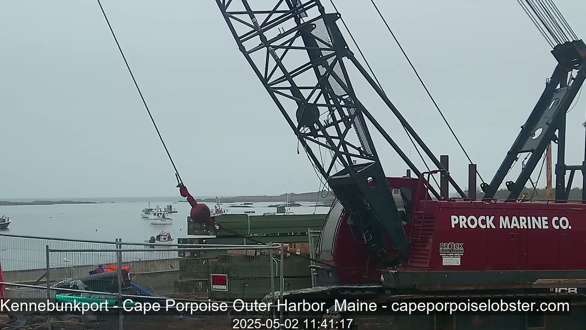 A red Prock Marine Co. crane lifts a large, dark green object near a harbor on an overcast day.